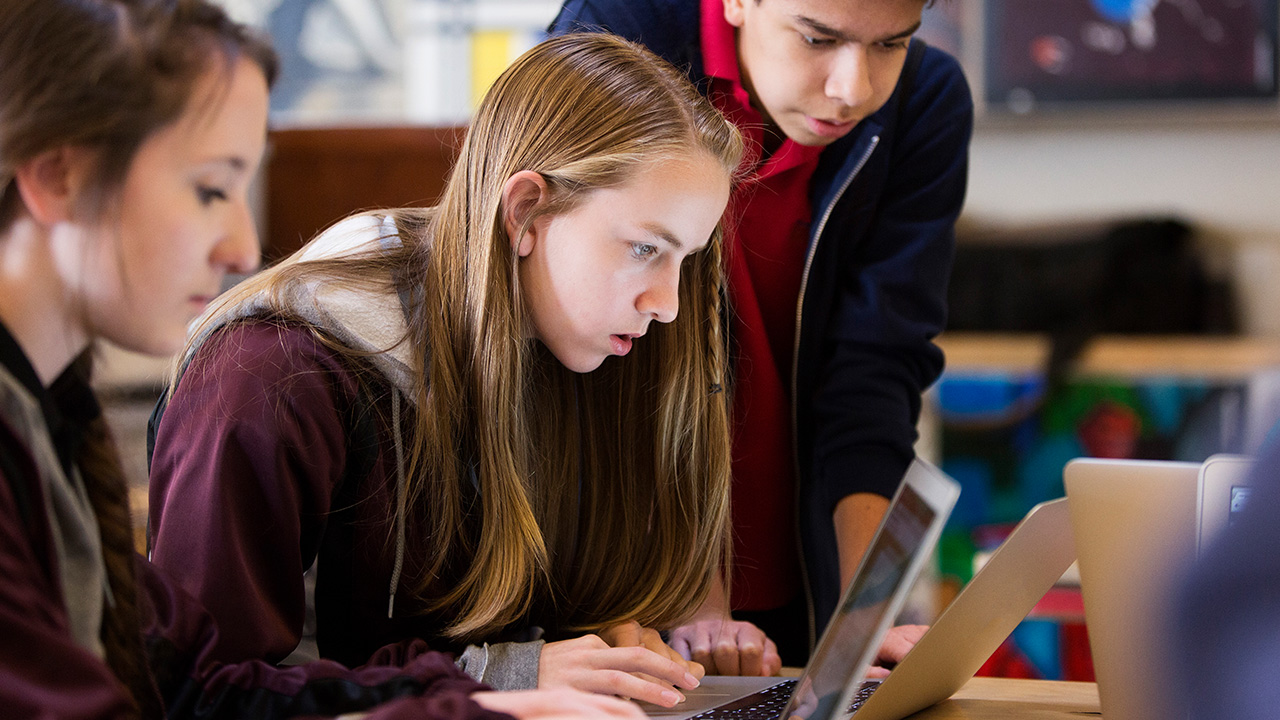 An image of a teen girl using her laptop with two others looking over her shoulder